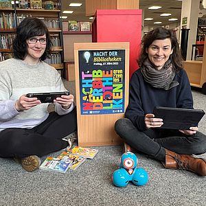 Zwei Frauchen sitzen in der Bibliothek. Die linke hält eine Spielkonsole in der Hand und die rechte ein Tablet. Im Hintergrund sind Regale zu sehen. Zwischen den beiden Frauen hängt das Plakat: Nacht der Bibliotheken